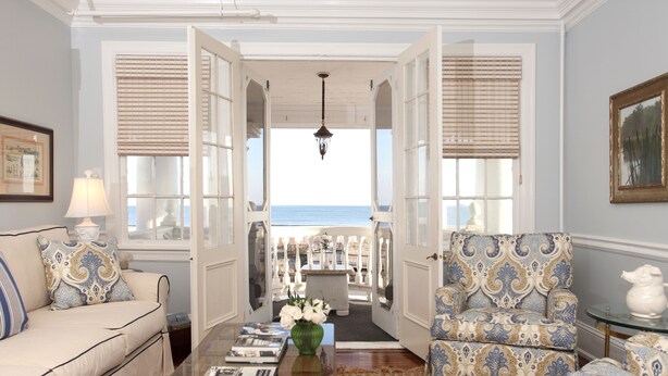 Una habitación con vista al mar, un sofá, un sillón estampado, una mesa de centro con libros y un jarrón con flores.