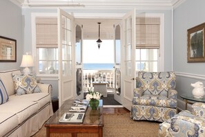 Una habitación con vista al mar, un sofá, un sillón estampado, una mesa de centro con libros y un jarrón con flores.