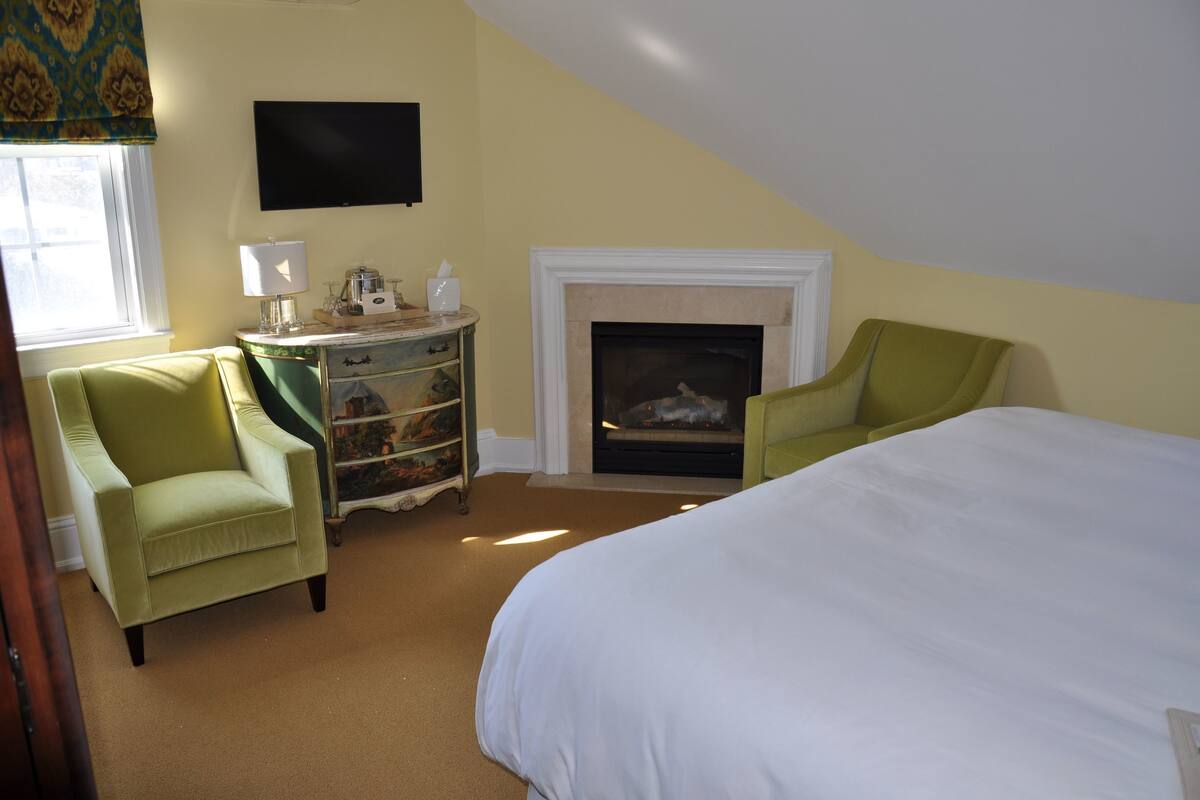 A bedroom with a bed, two chairs, a dresser, a television, and a fireplace.