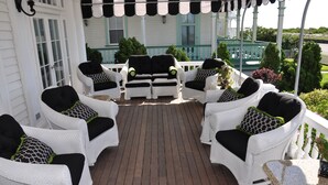 Un porche cubierto con mobiliario de mimbre blanco y cojines negros, que incluye una terraza de madera y vistas a un edificio blanco con detalles verdes.