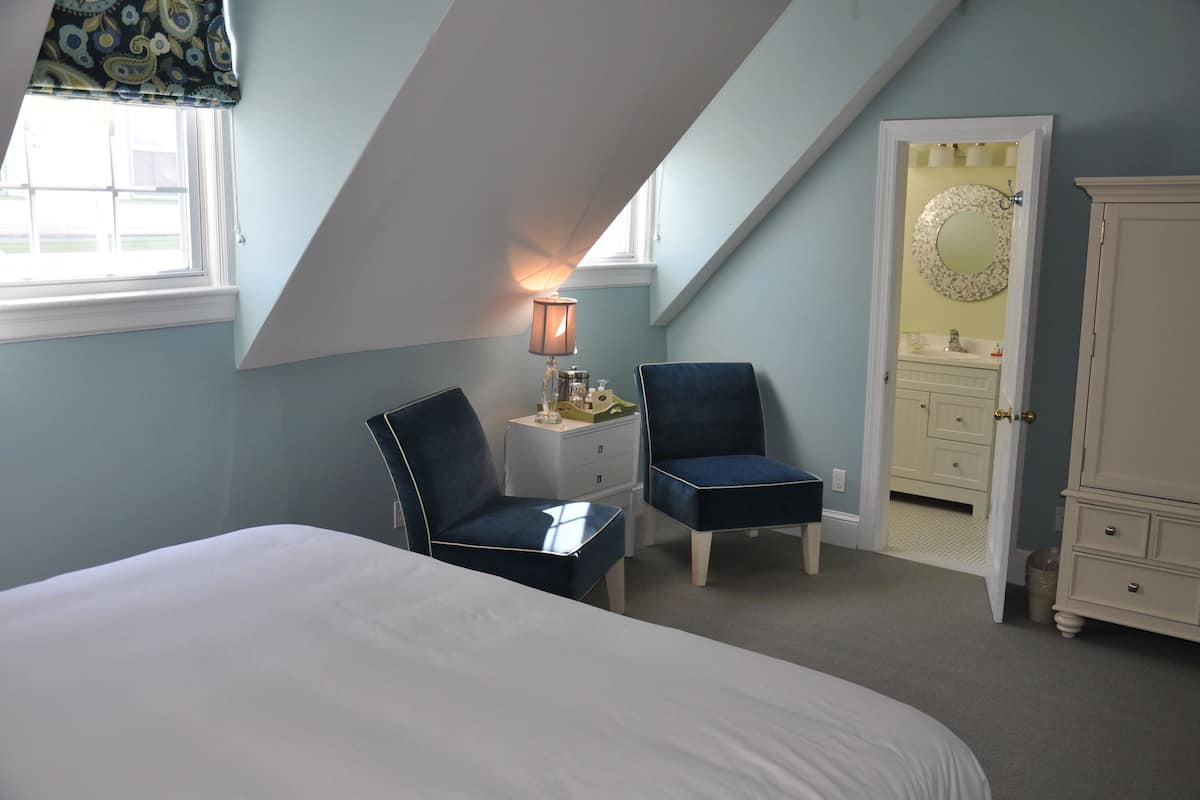 A bedroom with a bed, two chairs, a nightstand, a lamp, a dresser, and a door leading to a bathroom.