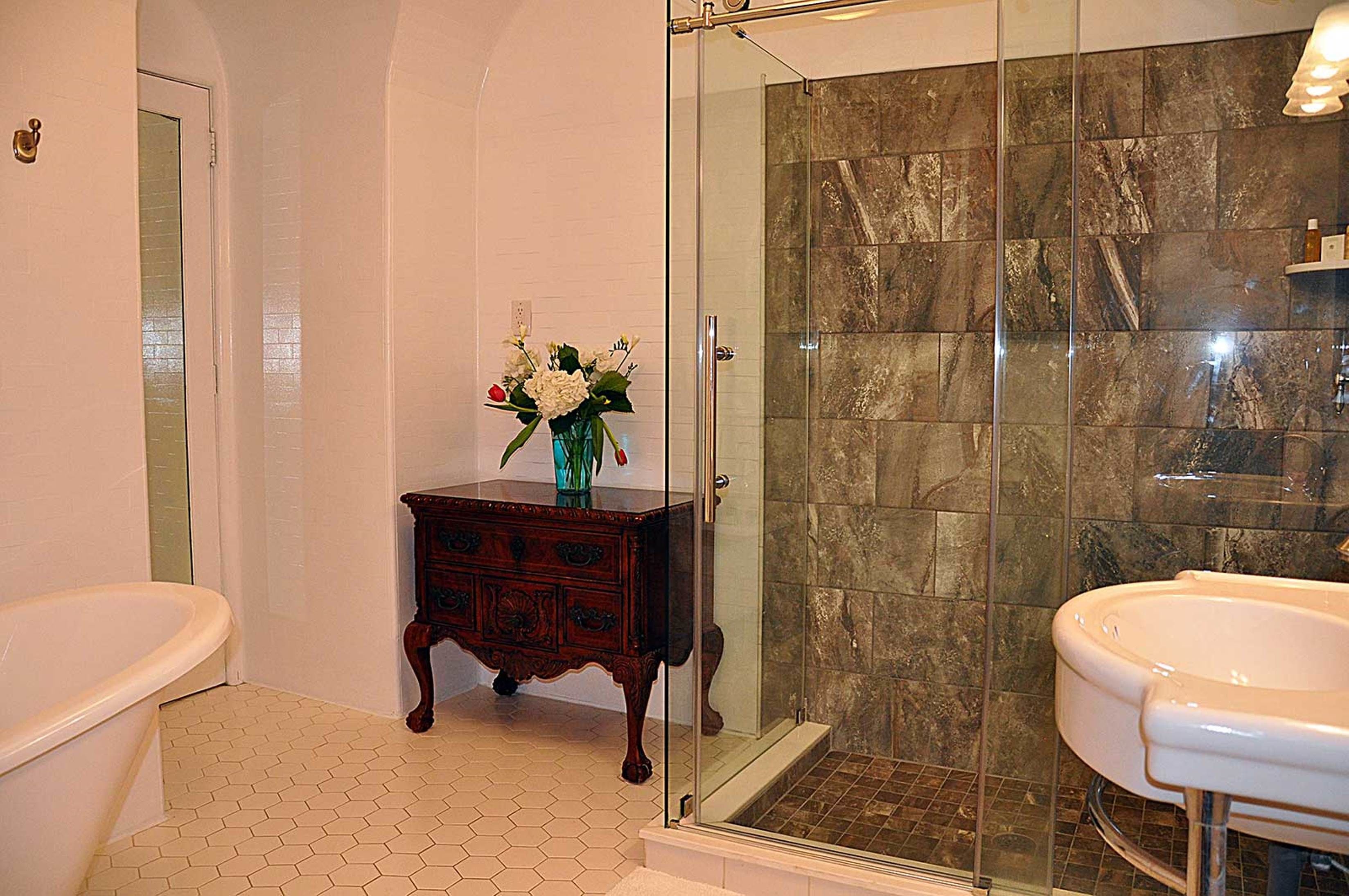A bathroom with a glass-enclosed shower, a white bathtub, and a wooden vanity with a vase of flowers.