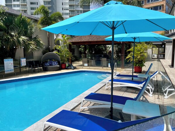 A swimming pool with blue lounge chairs and umbrellas, surrounded by palm trees and a building in the background. - Burleigh Palms Holiday Apartments (Burleigh Heads)