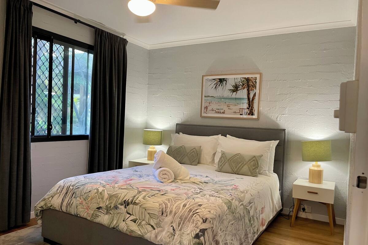 A bedroom with a bed, two bedside tables with lamps, a window with curtains, a ceiling fan, and a beach-themed wall art.