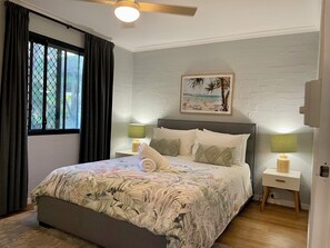 A bedroom with a bed, two bedside tables with lamps, a window with curtains, a ceiling fan, and a beach-themed wall art. - Burleigh Palms Holiday Apartments (Burleigh Heads)