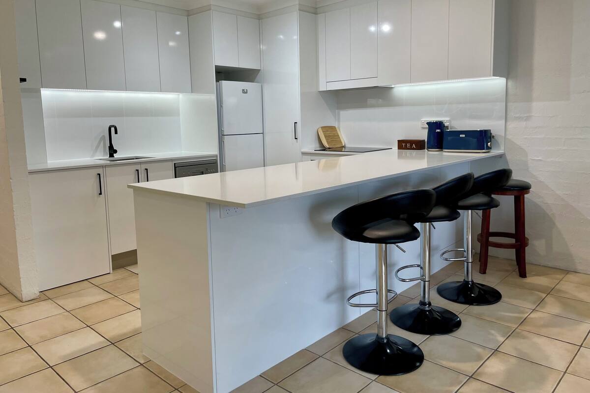 A modern kitchen with white cabinetry, a central island with bar stools, a built-in dishwasher, and a coffee machine.