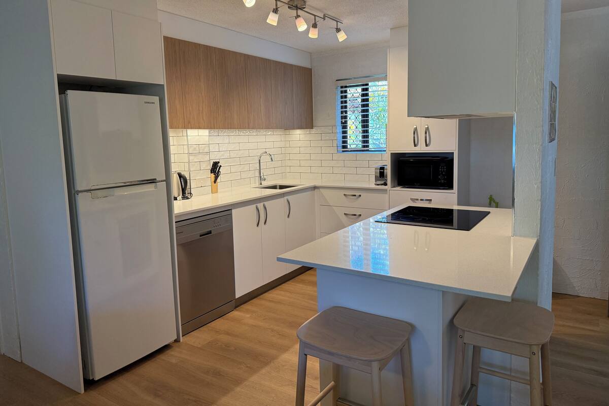 A modern kitchen with white cabinetry, a built-in dishwasher, a stainless steel refrigerator, a microwave, and a large island with stools.