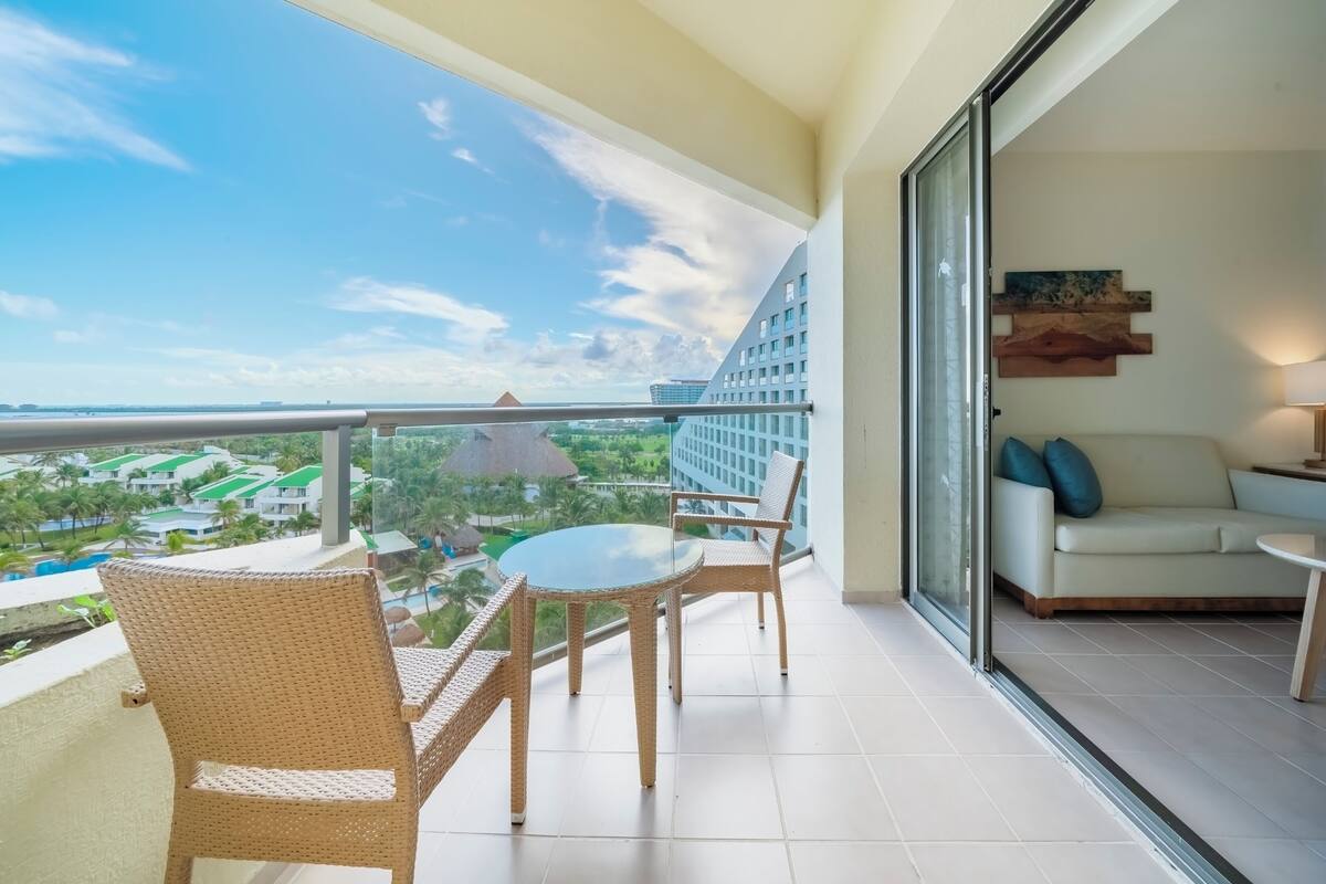 Un balcón con mobiliario de mimbre, un ventanal de vidrio que da a una sala de estar con un sofá y una lámpara, y vistas a un resort con edificios y palmeras.