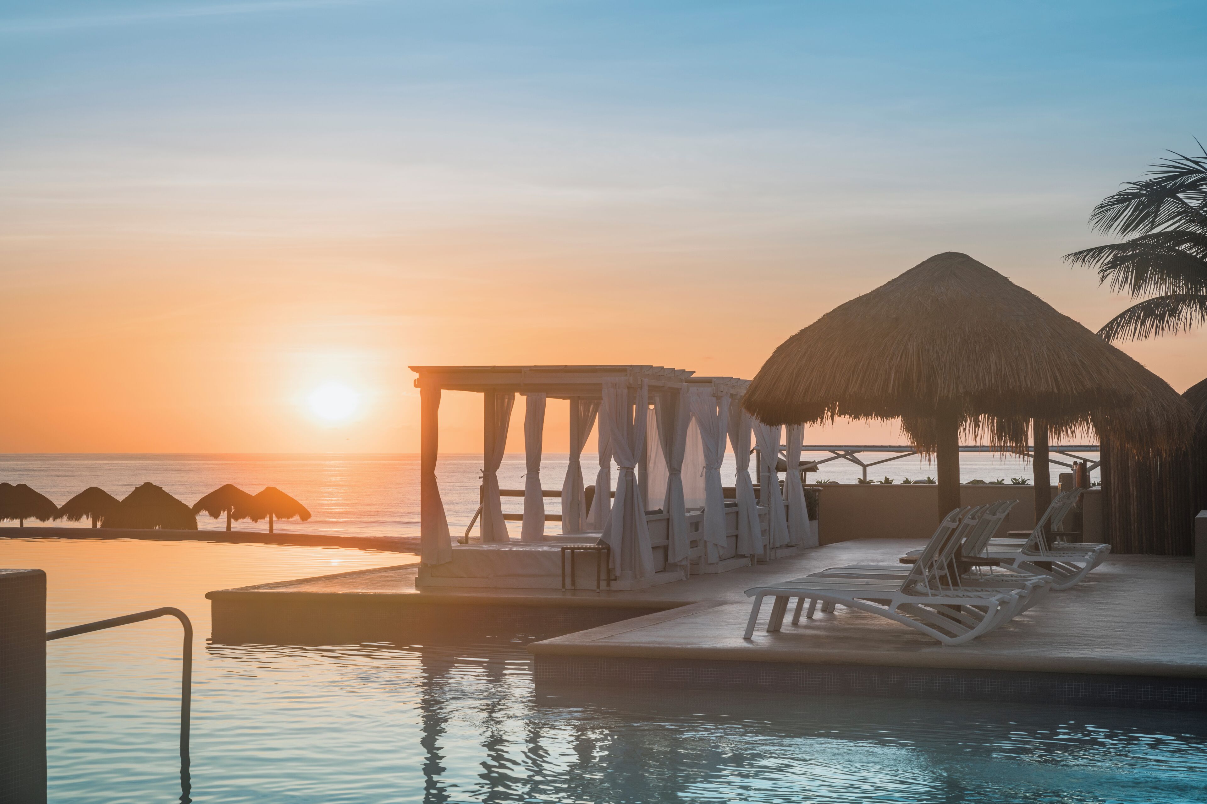 A resort pool area with a thatched-roof structure, lounge chairs, and a view of the sunset over the ocean.