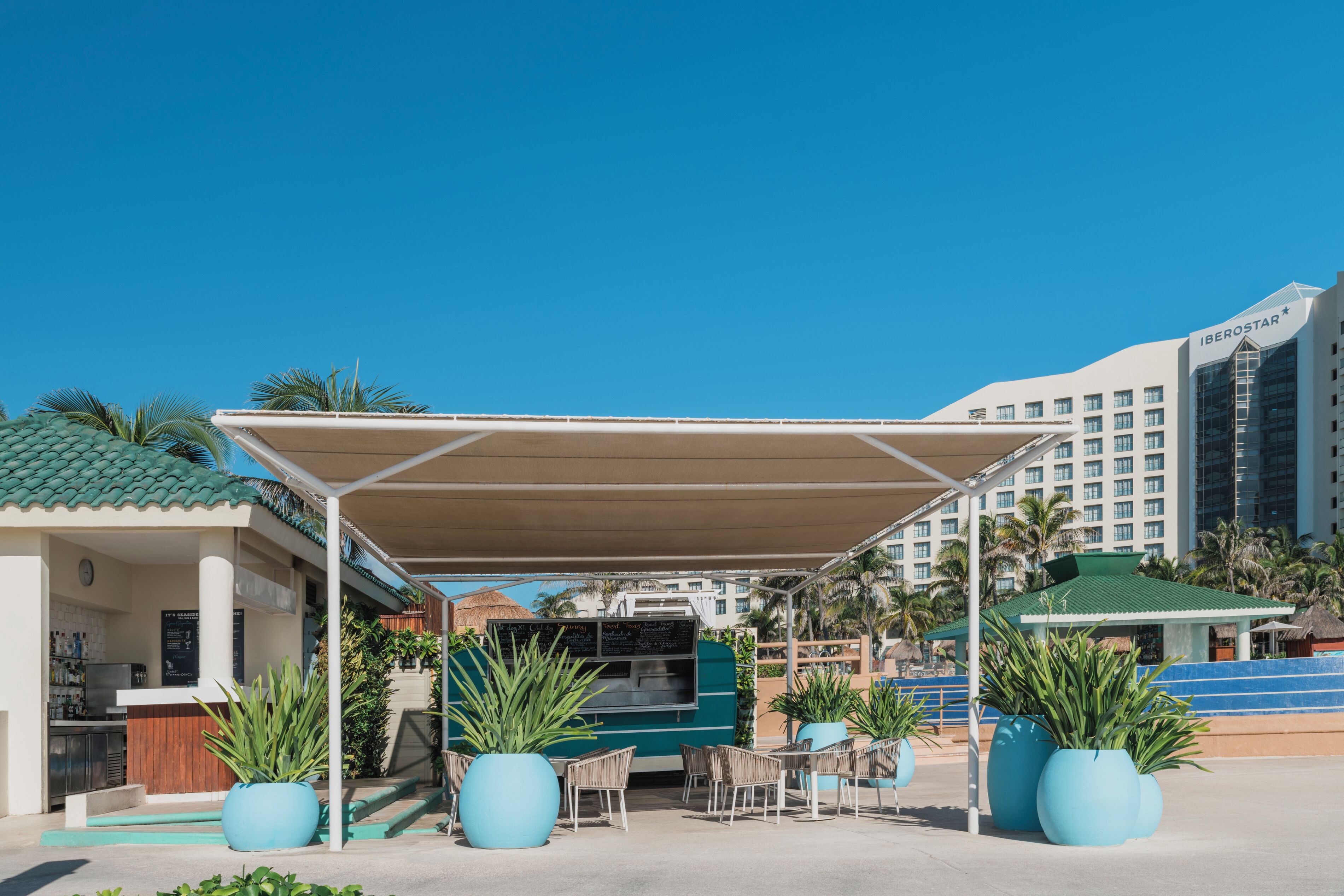 A covered outdoor seating area with large planters, a food kiosk, and a view of hotel buildings.