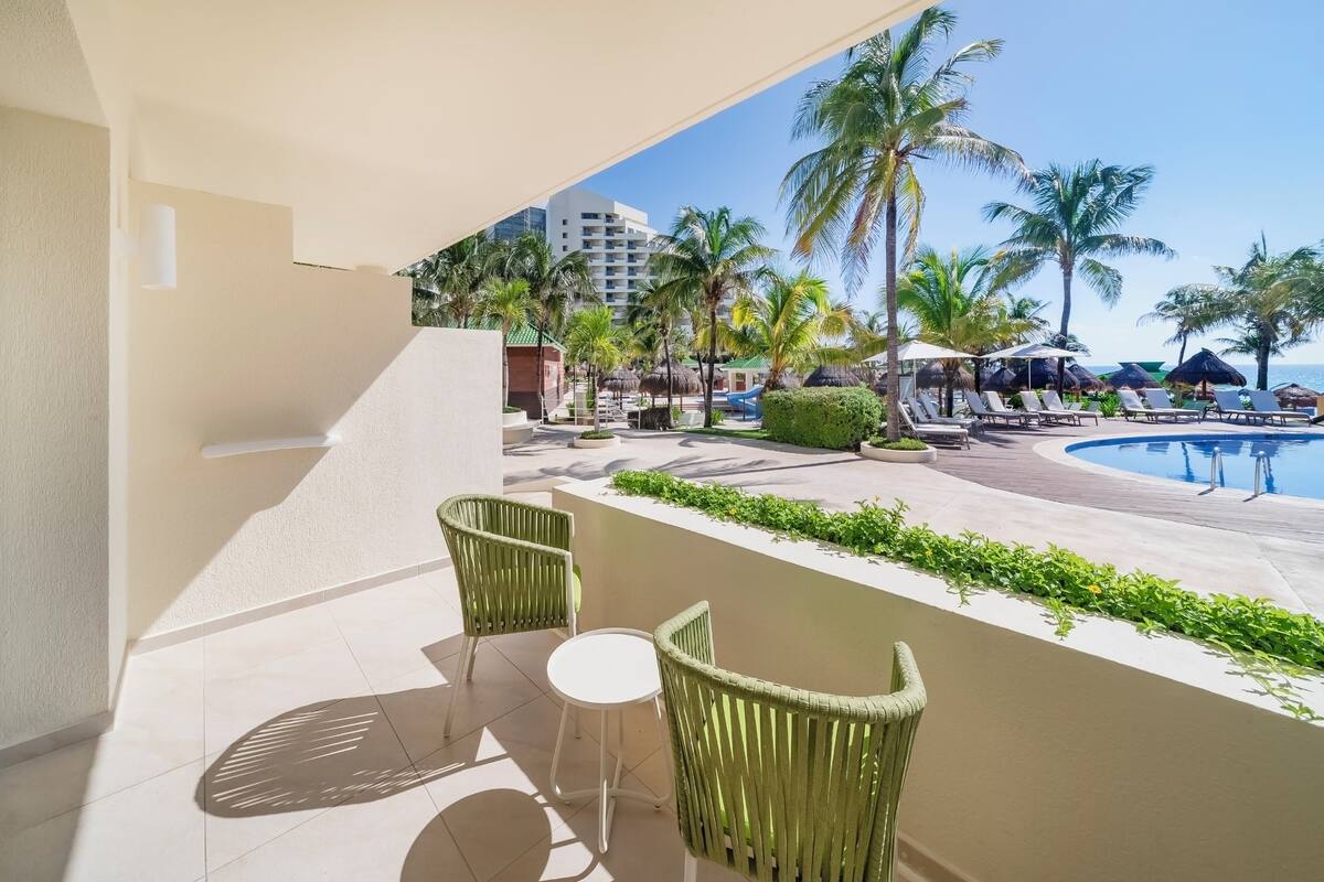 Un balcón con sillas de mimbre y una pequeña mesa, con vistas a una zona de piscina con sillones de descanso y palmeras.