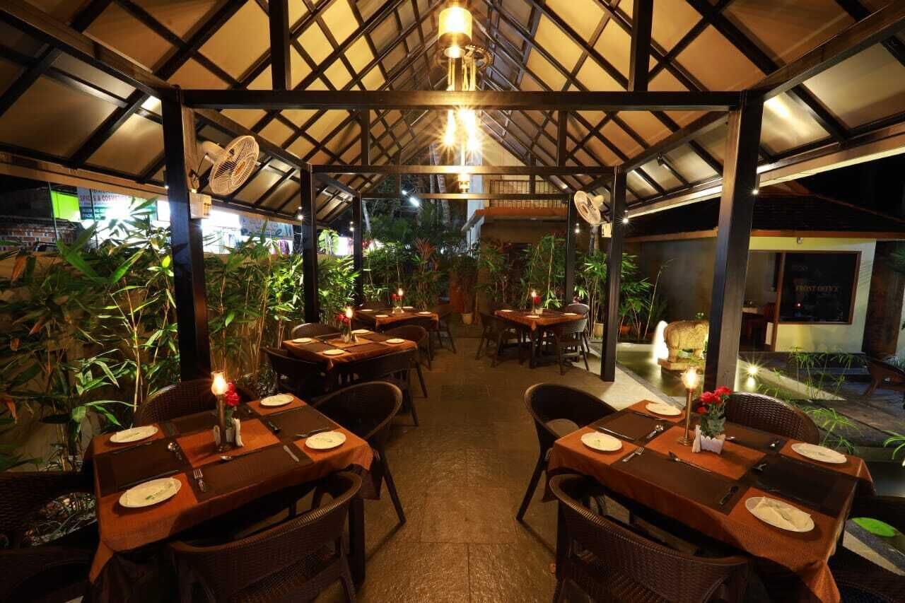 Un comedor interior con mesas dispuestas para dos personas, rodeado de plantas y una estructura de techo de madera.