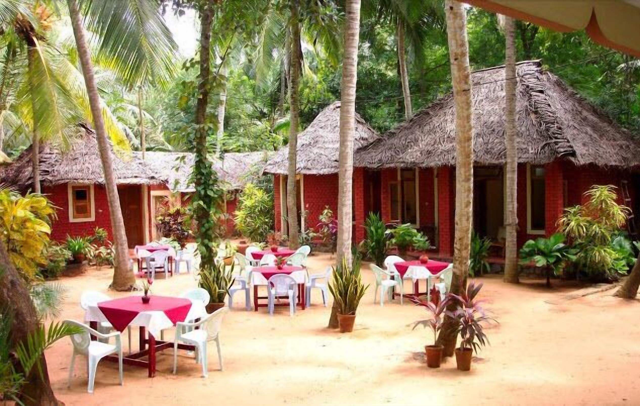 A tropical resort with thatched-roof huts, outdoor dining areas with white plastic chairs, and lush greenery.
