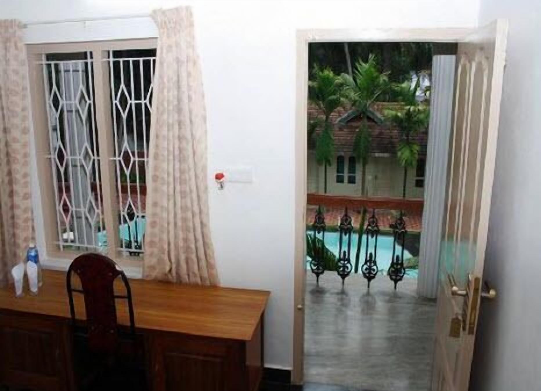 Una habitación con una mesa y sillas de madera, un ventanal con barrotes y cortinas, y una puerta que da a un área exterior con piscina y plantas.