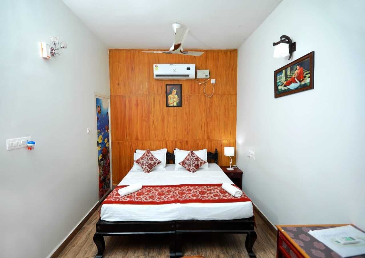 Une chambre d’hôtel avec une tête de lit en bois, un lit avec une literie blanche et rouge, un appareil de climatisation, une lampe de chevet et un tableau encadré au mur.