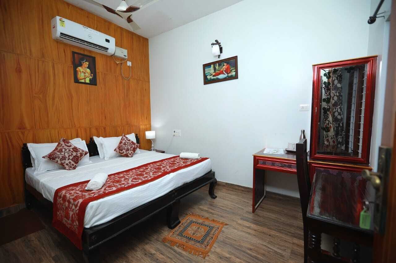 Habitación de hotel con cabecera de madera, cama con ropa blanca y detalles rojos, aire acondicionado, una mesita con lámpara y un espejo con marco decorativo.