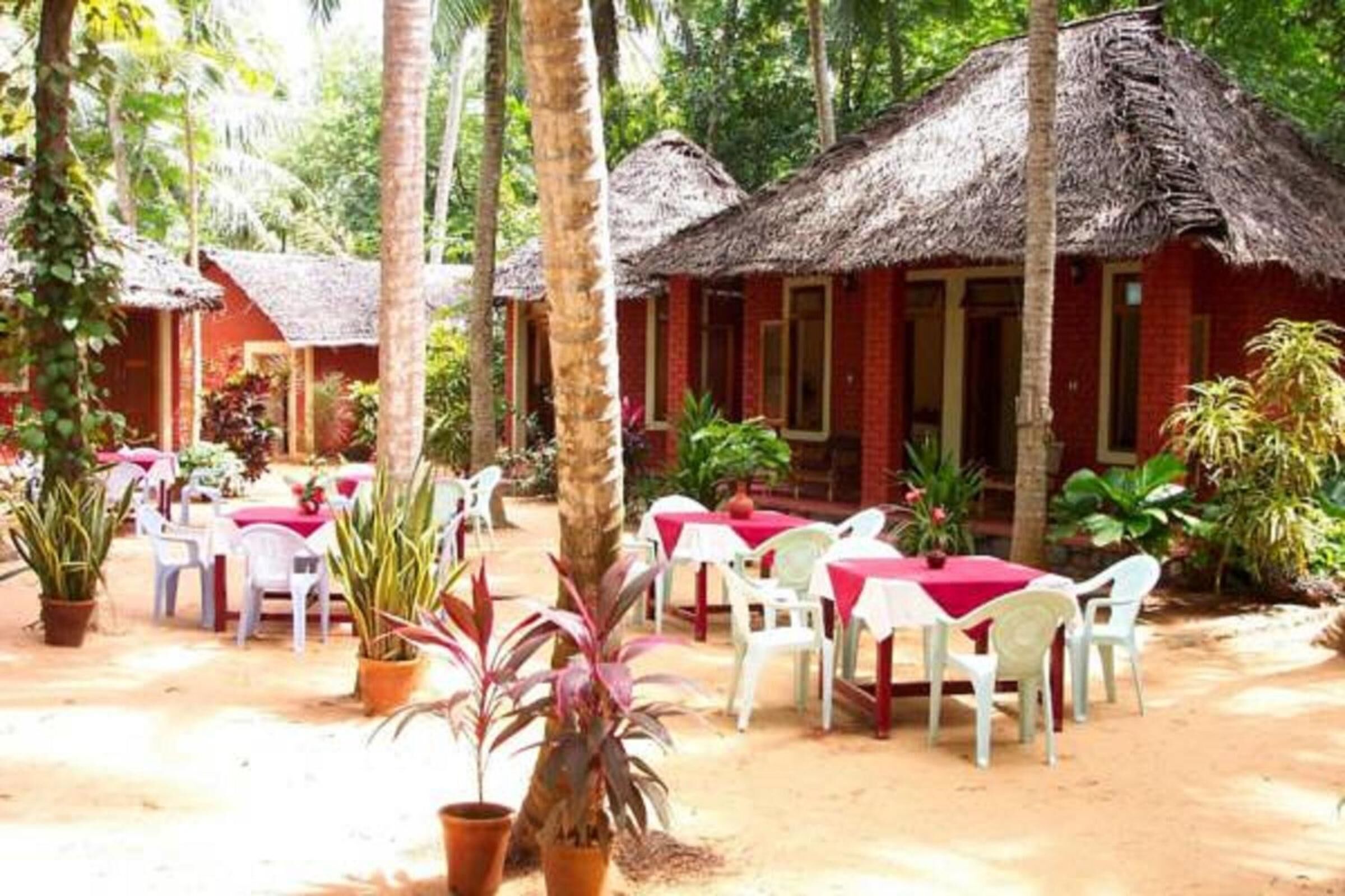 A resort with thatched-roof huts, outdoor dining areas with white plastic chairs and red tablecloths, surrounded by potted plants and palm trees.