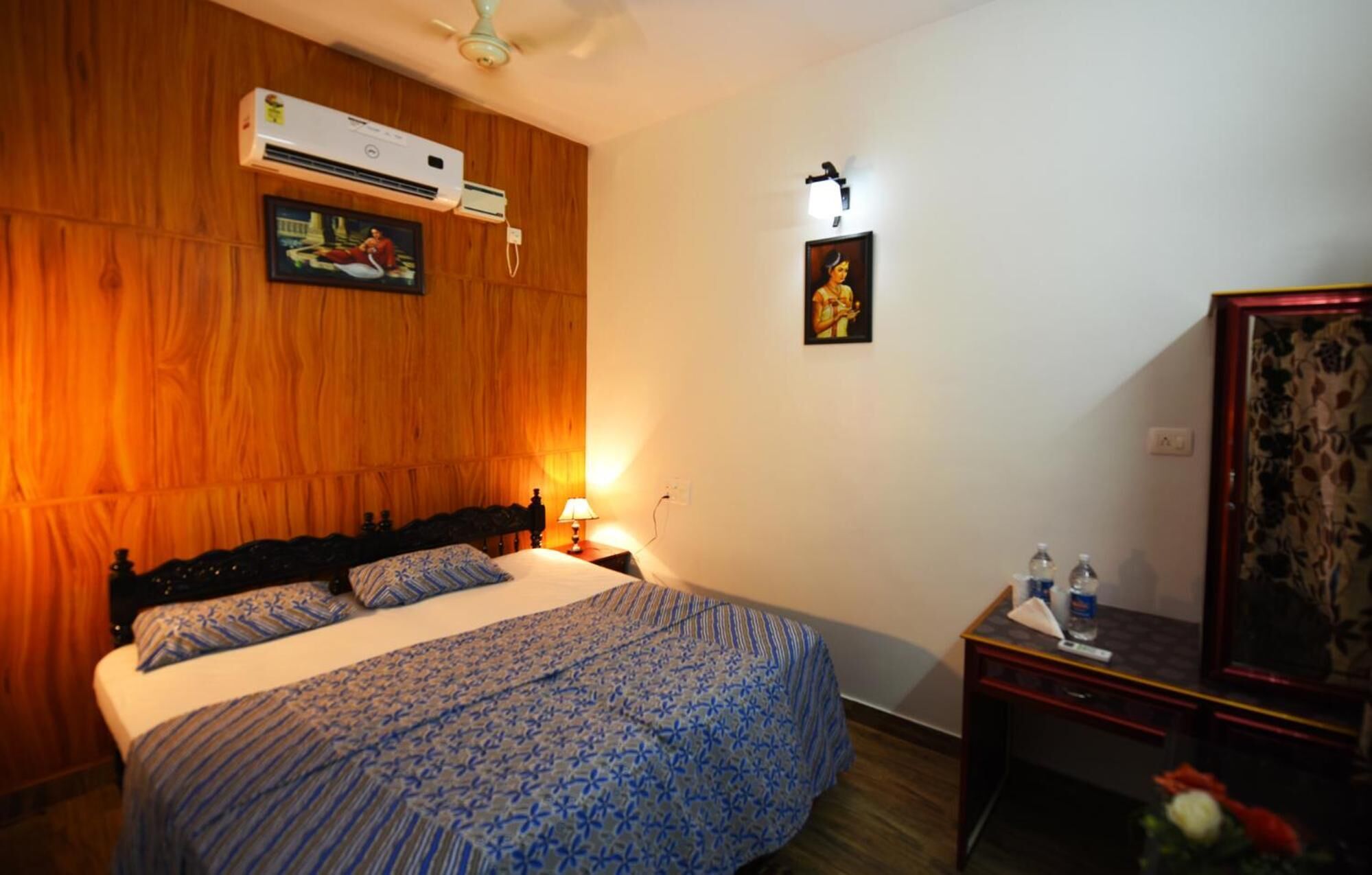 Een slaapkamer met een houten hoofdeinde, een bed met een blauw gebedrukte spreideken, een airconditioning, een nachtkastje met een lamp en een aan de muur hangend schilderij.