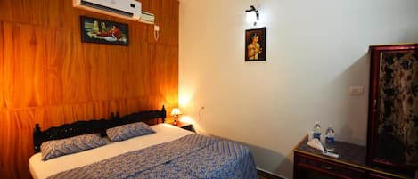 Un dormitorio con cabecera de madera, cama con colcha azul estampada, unidad de aire acondicionado, mesita de noche con lámpara y un cuadro colgado en la pared.