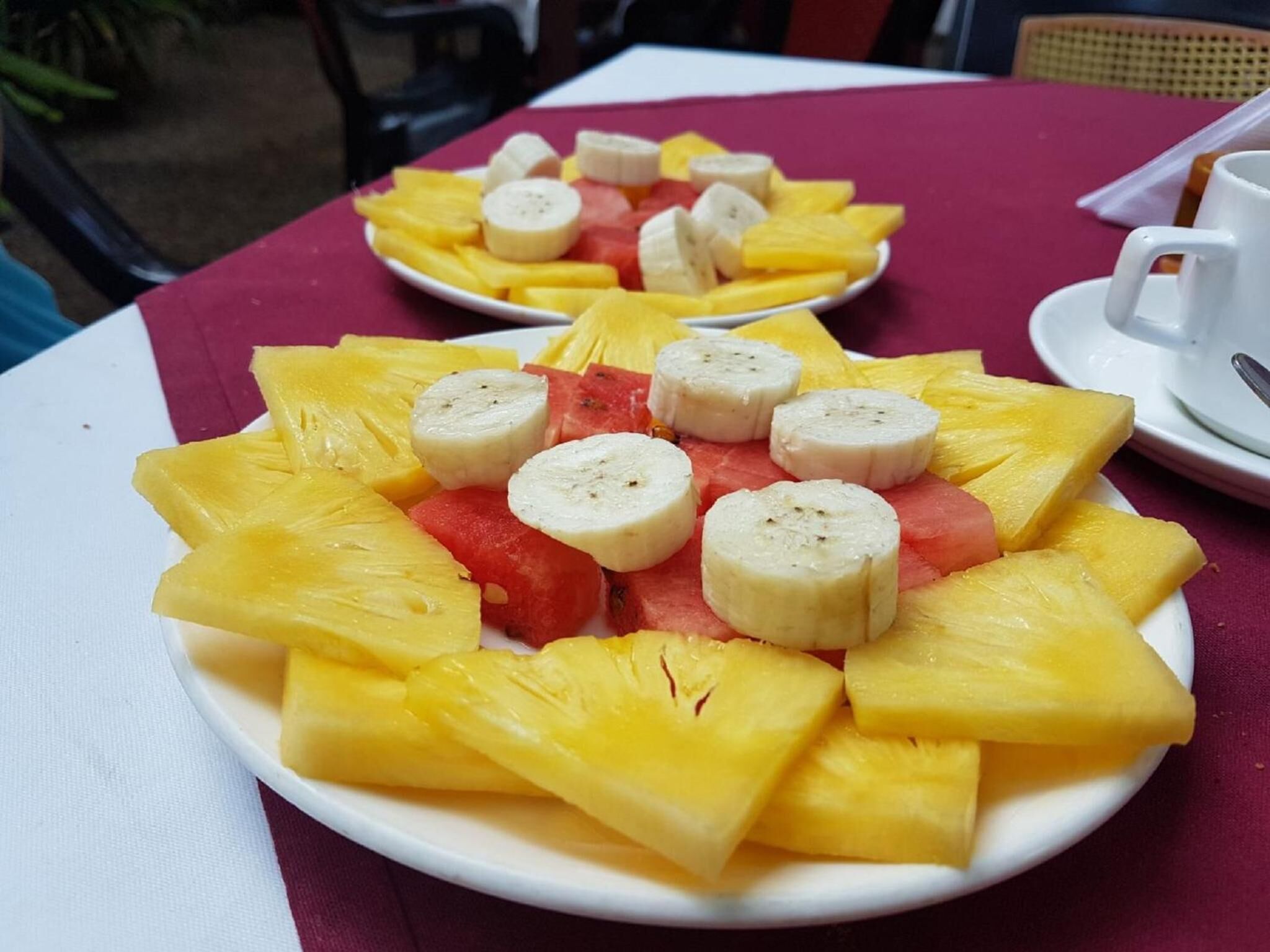 Ett bord med en vit tallrik fylld med en fruktsallad som består av ananas, vattenmelon och bananskivor.