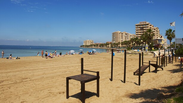 Una playa con arena, sombrillas y gente. Se ven edificios al fondo.