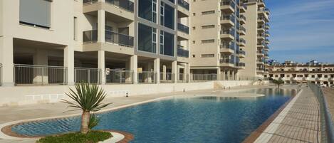 A modern hotel with a swimming pool, multiple balconies, and a clear sky.