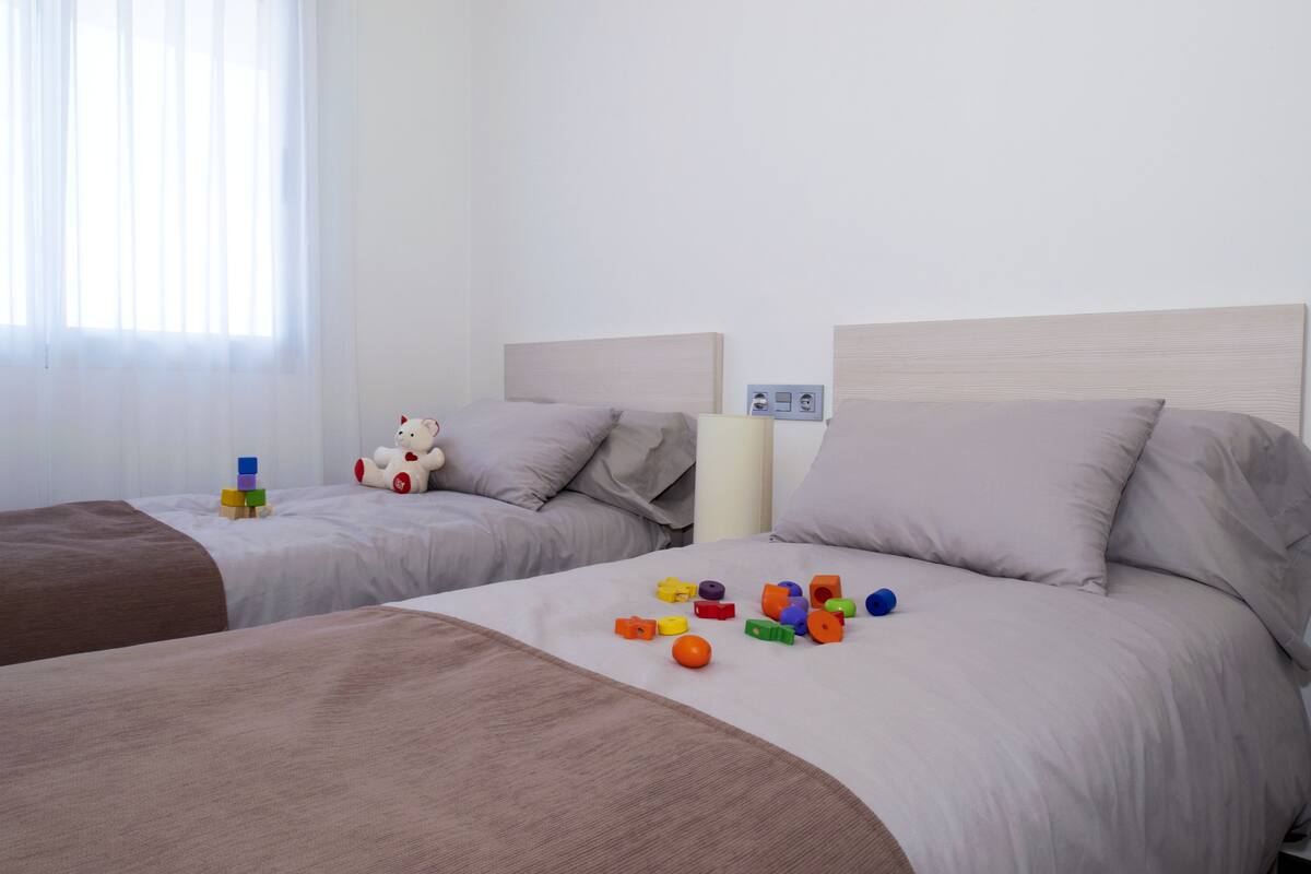 A hotel room with two beds, each with a headboard and pillows. There are toys on the beds and a window with curtains.