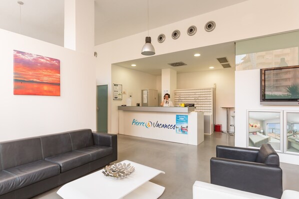 Un área de recepción moderna con un sofá gris, una mesa de centro blanca y un mostrador de recepción con la leyenda "Pierre & Vacances".