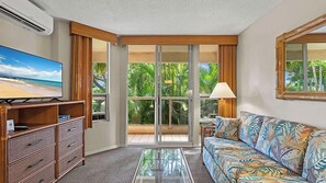 A hotel room with a television, a sofa, a coffee table, a dresser, and a sliding glass door leading to a balcony with a view of palm trees. - Maui Banyan Vacation Club (Kihei)