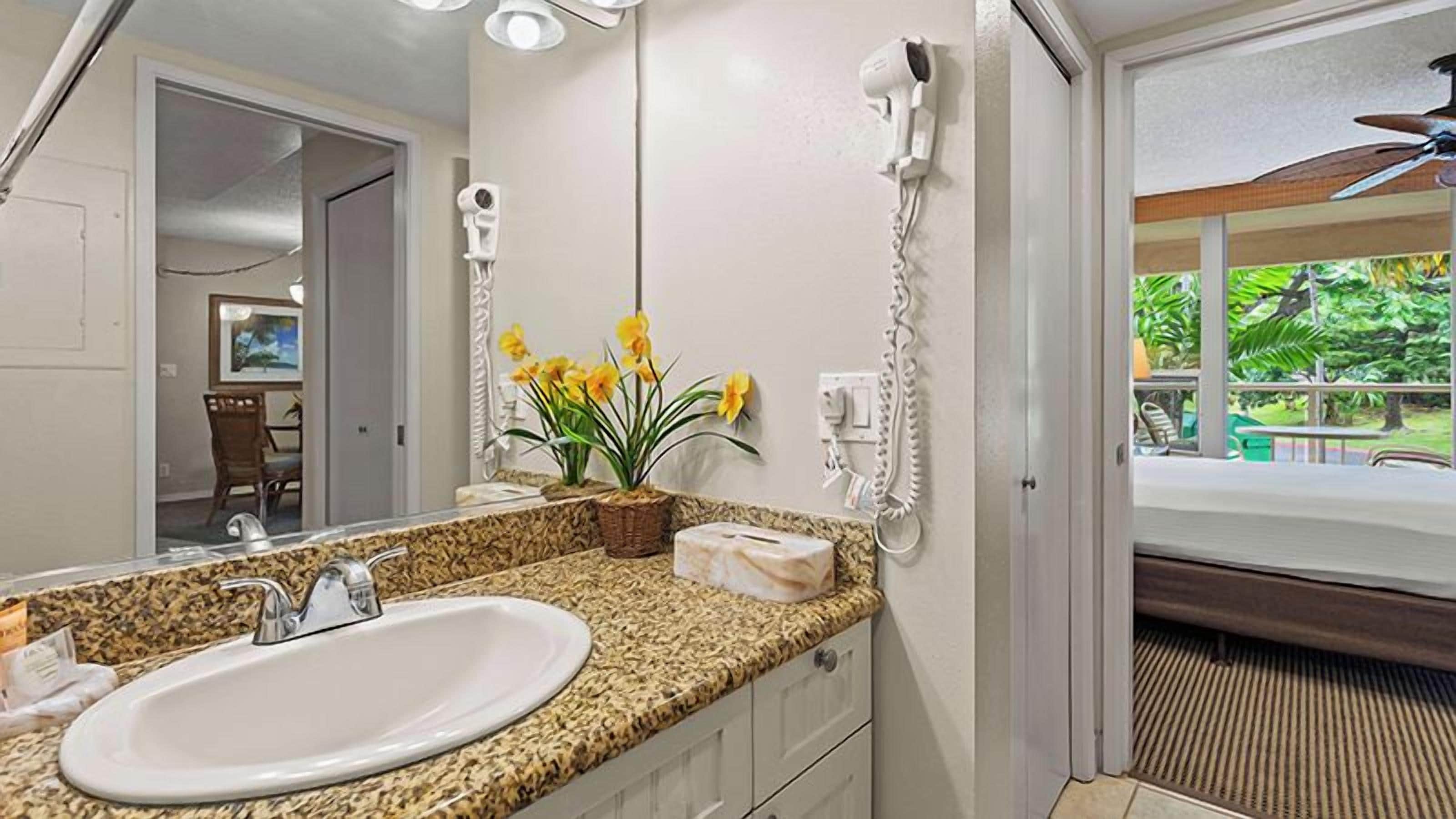 A bathroom with a granite countertop sink, a hairdryer, and a window with a view of greenery.