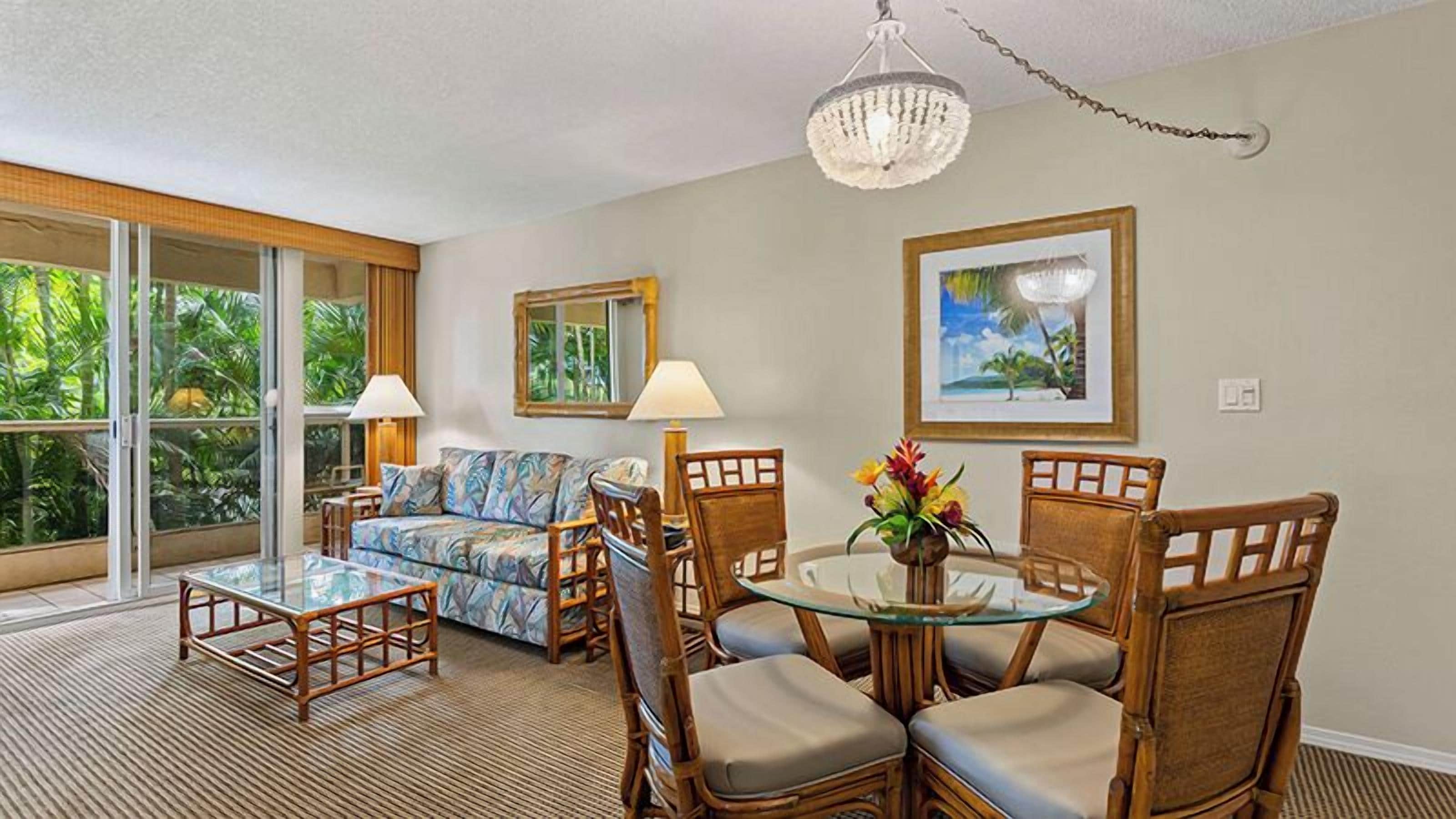 A living room with a sofa, coffee table, and dining table with chairs. There is a large window with a view of greenery.