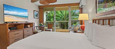 A hotel room with a large bed, a television, a desk with a chair, a window with a view of palm trees, and a ceiling fan.