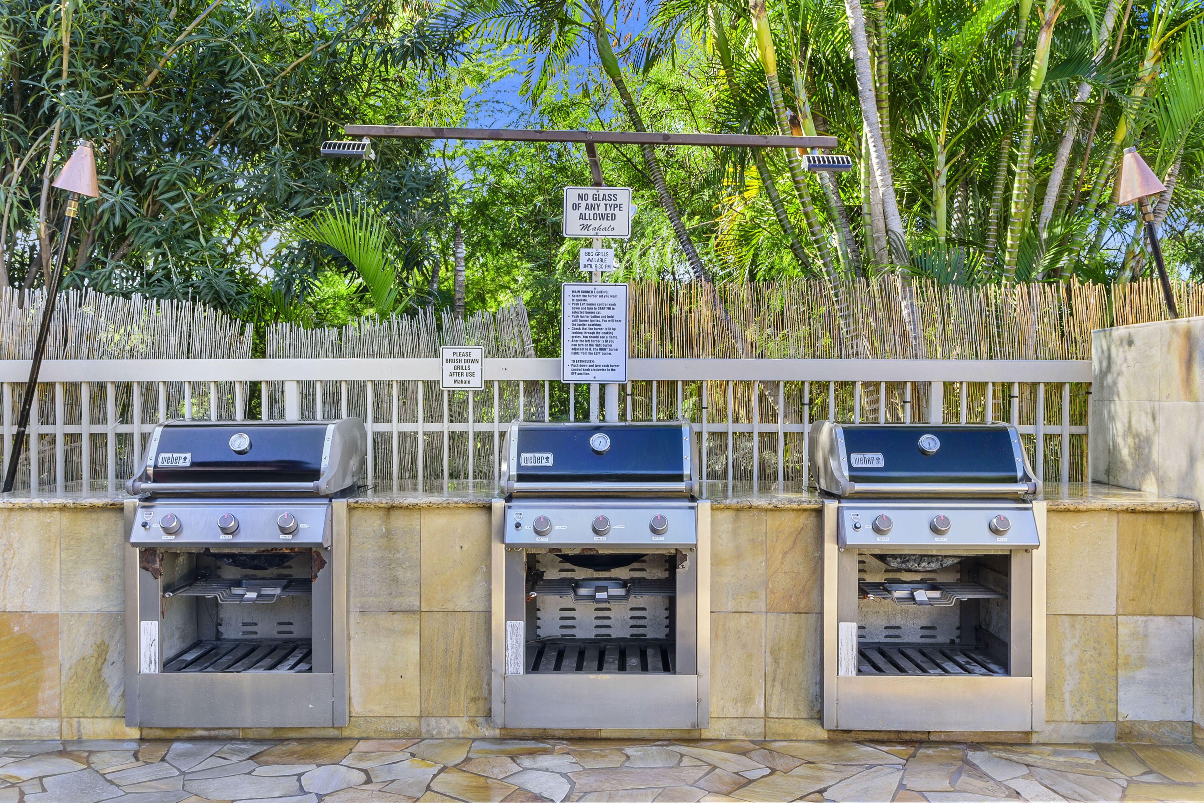 Trois barbecues sont placés devant une clôture en bambou, et un panneau indique qu’il est interdit d’utiliser des récipients en verre.