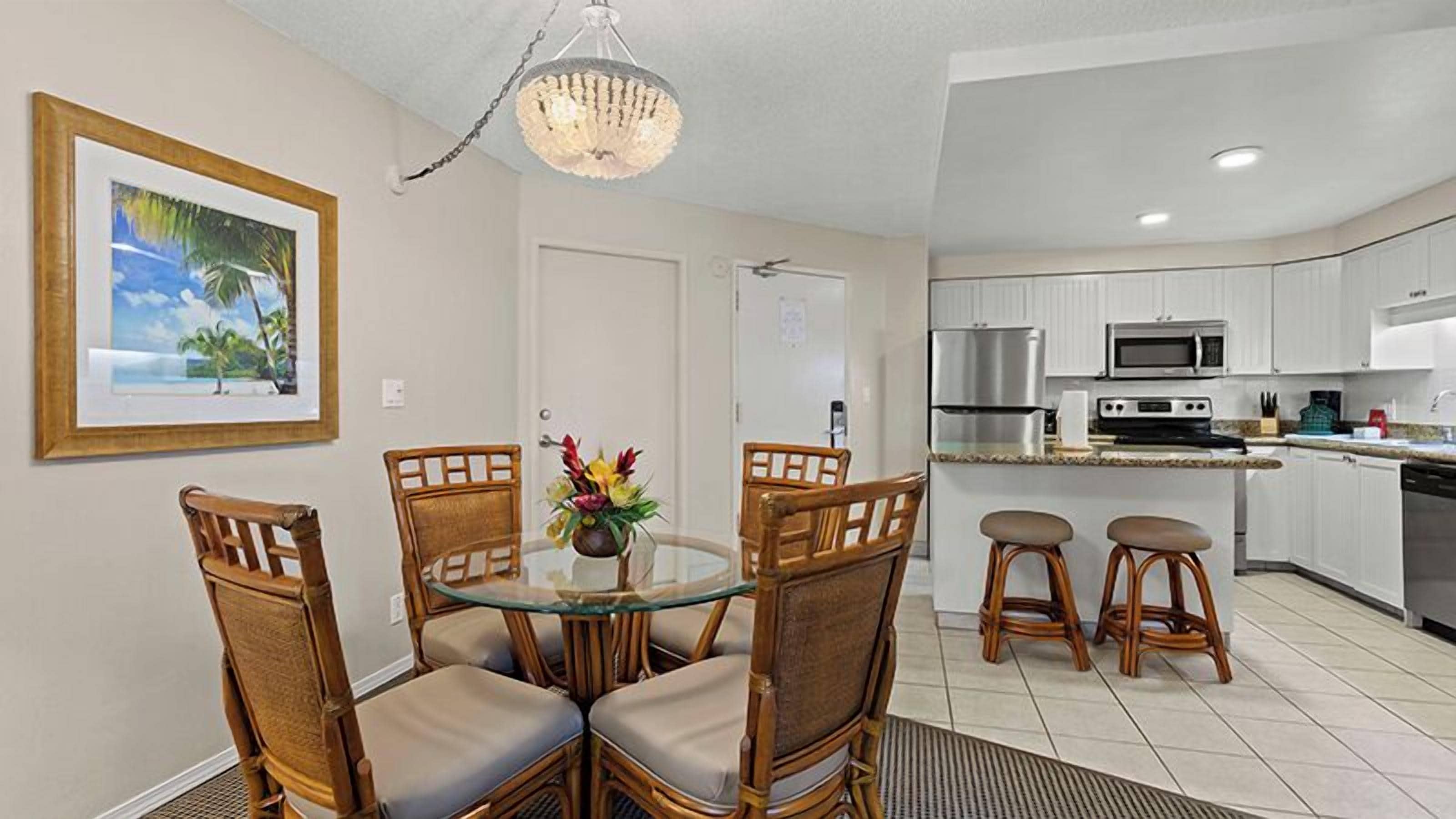 A dining area with a glass table and wicker chairs, a kitchen with white cabinets and stainless steel appliances, and a framed picture on the wall.