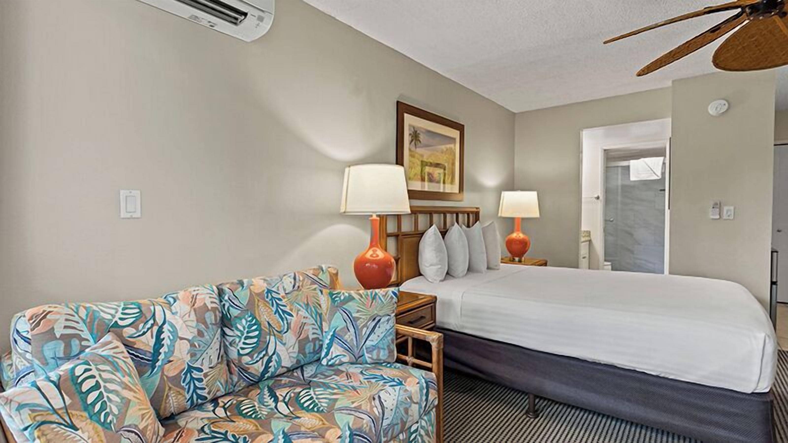 A hotel room with a bed, a sofa, two lamps, a ceiling fan, and a door.