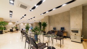 A modern hotel lobby with a glass-top table surrounded by wicker chairs, a reception desk with computers, and several potted plants. - Reborn Suwon Silkroad Hotel (Suwon)
