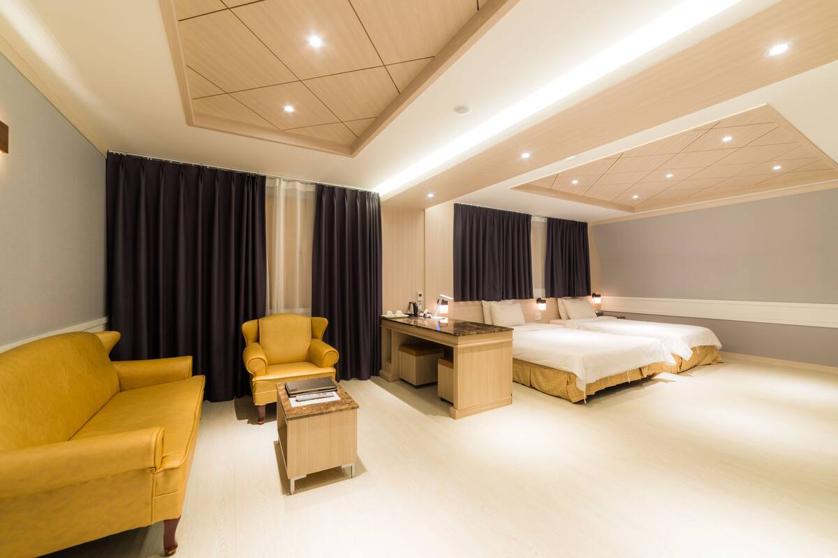 A modern hotel room with a yellow sofa, a desk with a computer, a bed, and a large mirror.