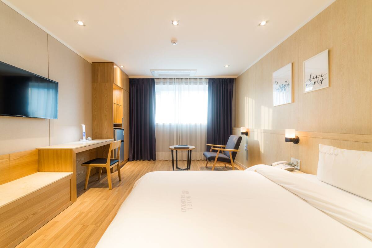 A modern hotel room with a large bed, a desk, a chair, a small table, and a TV mounted on the wall.