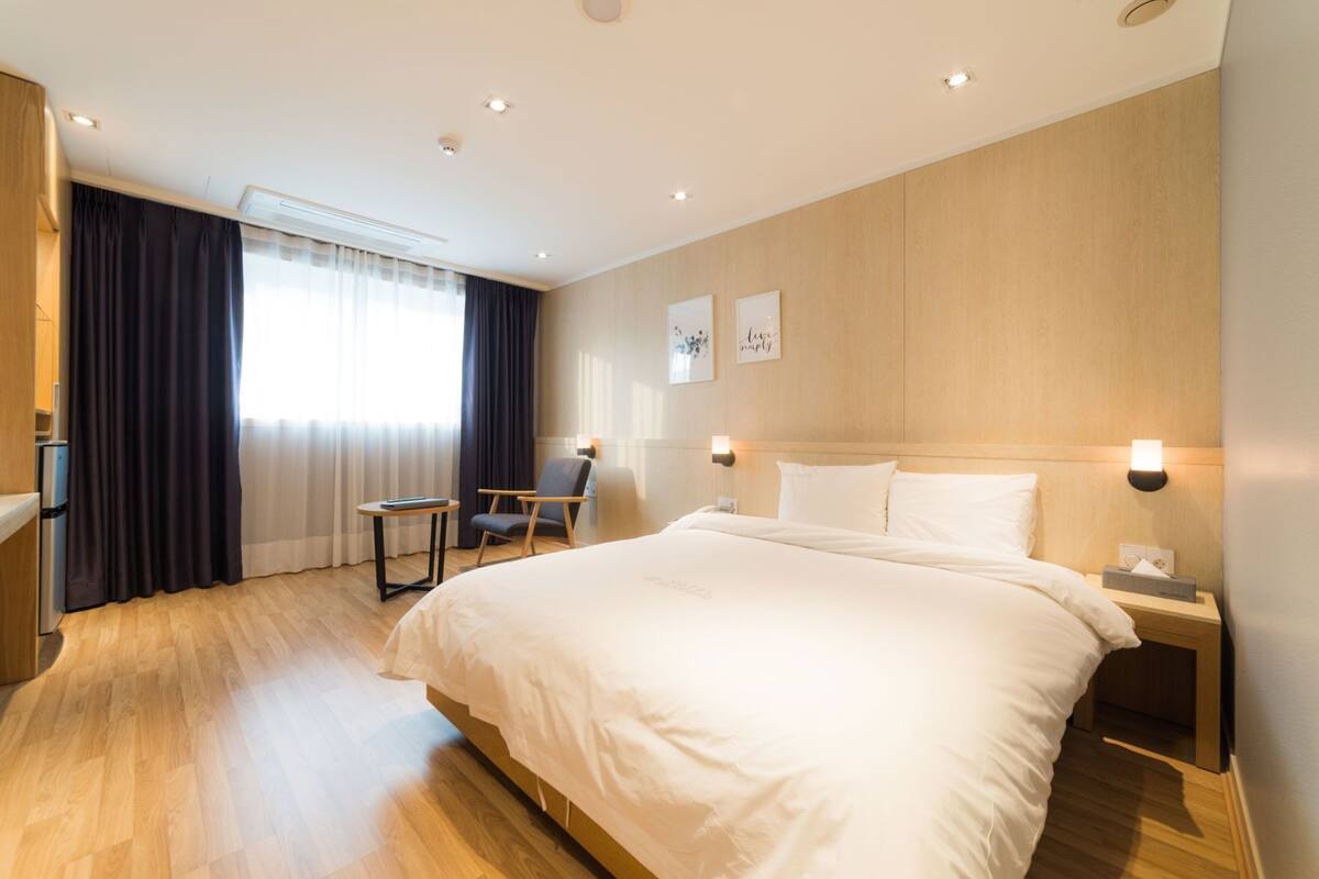 A hotel room with a large bed, a desk, a chair, a window with curtains, and wall-mounted lights.