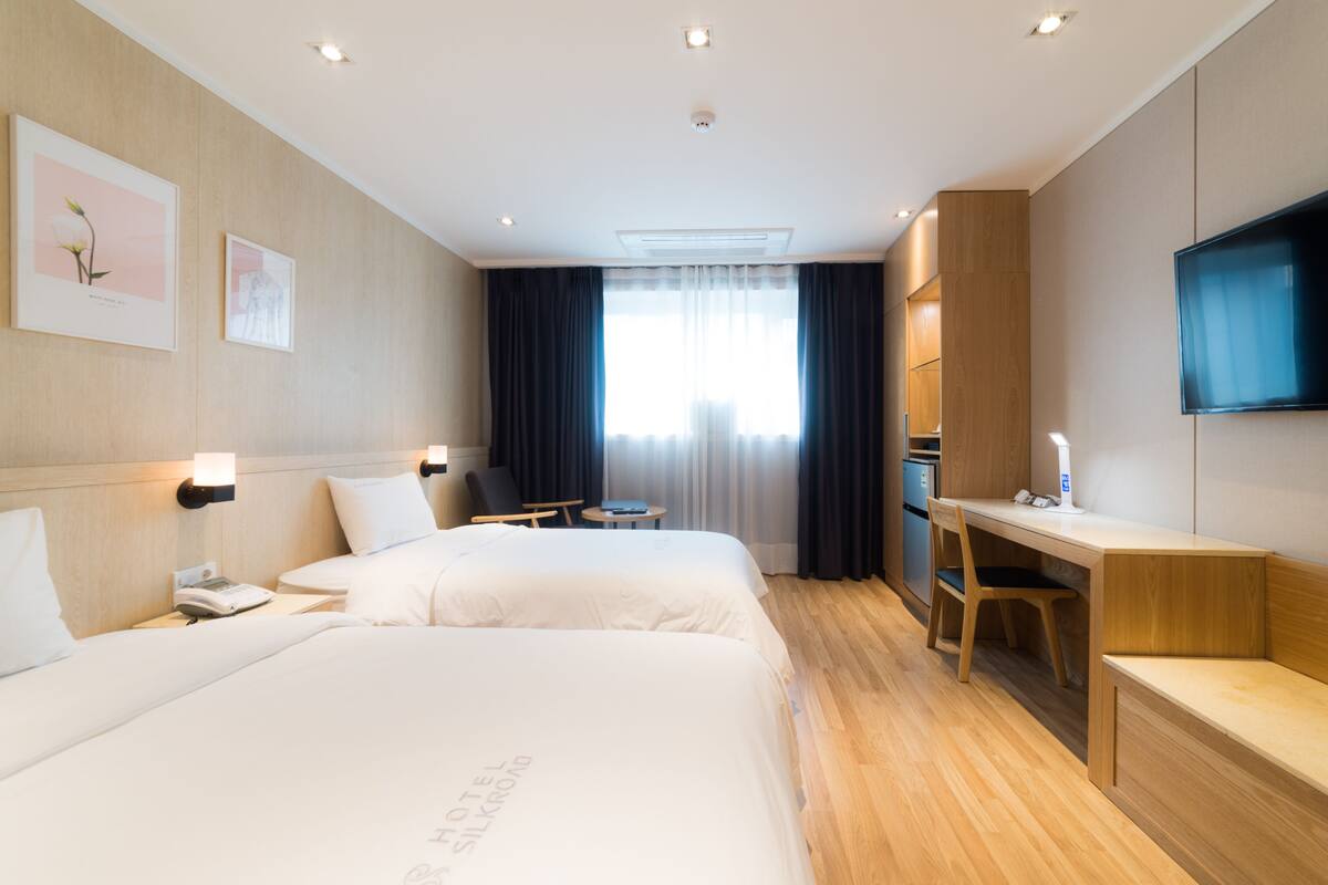 A hotel room with two beds, a desk, a chair, a TV, and a window with curtains.
