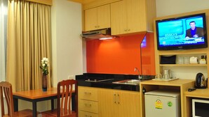 A compact kitchen with wooden cabinets, a black countertop, a small refrigerator, a TV displaying a show, and a dining area with a table and chairs. - Green Harbor Hotel & Service Apartment (Patong)
