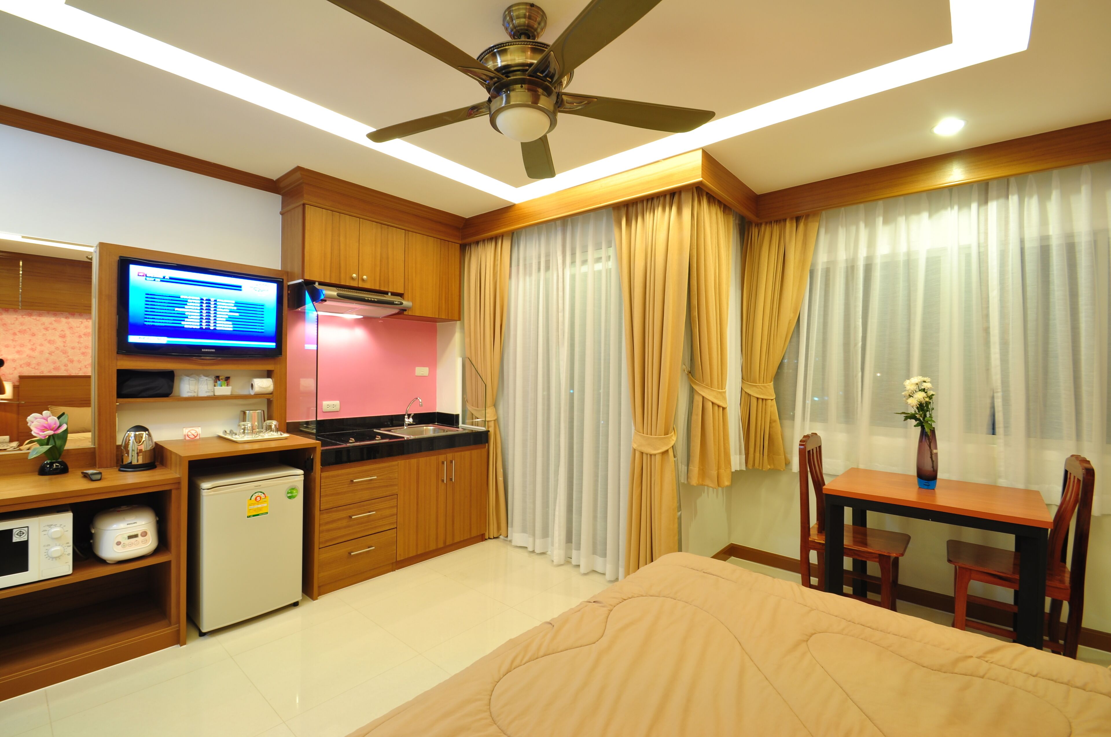 A compact hotel room with a kitchenette, a bed, a small table with chairs, and a ceiling fan.