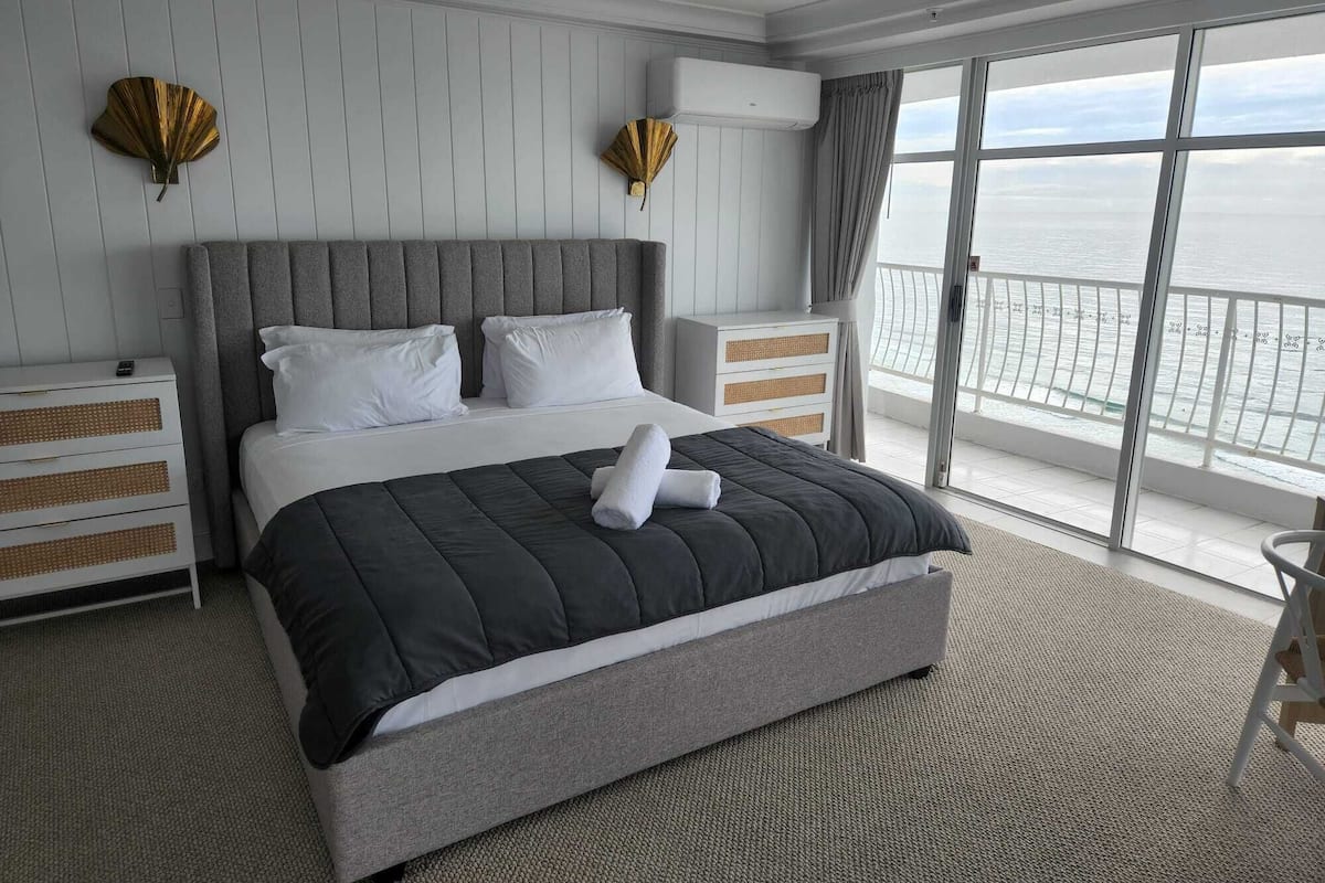 A modern hotel room with a large bed, a balcony with a view of the sea, and a white dresser with drawers.