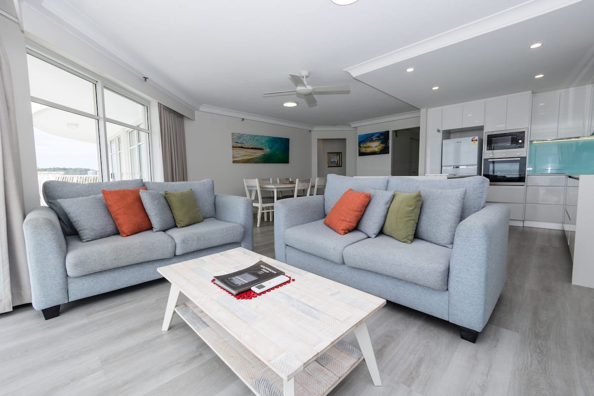 A modern living room with two grey sofas, a wooden coffee table, and a dining area with a white table and chairs.