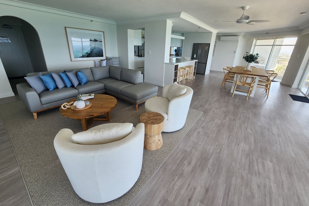 A spacious living room with a grey sectional sofa, a round coffee table, and a white armchair. A dining area with a wooden table and chairs is visible in the background.