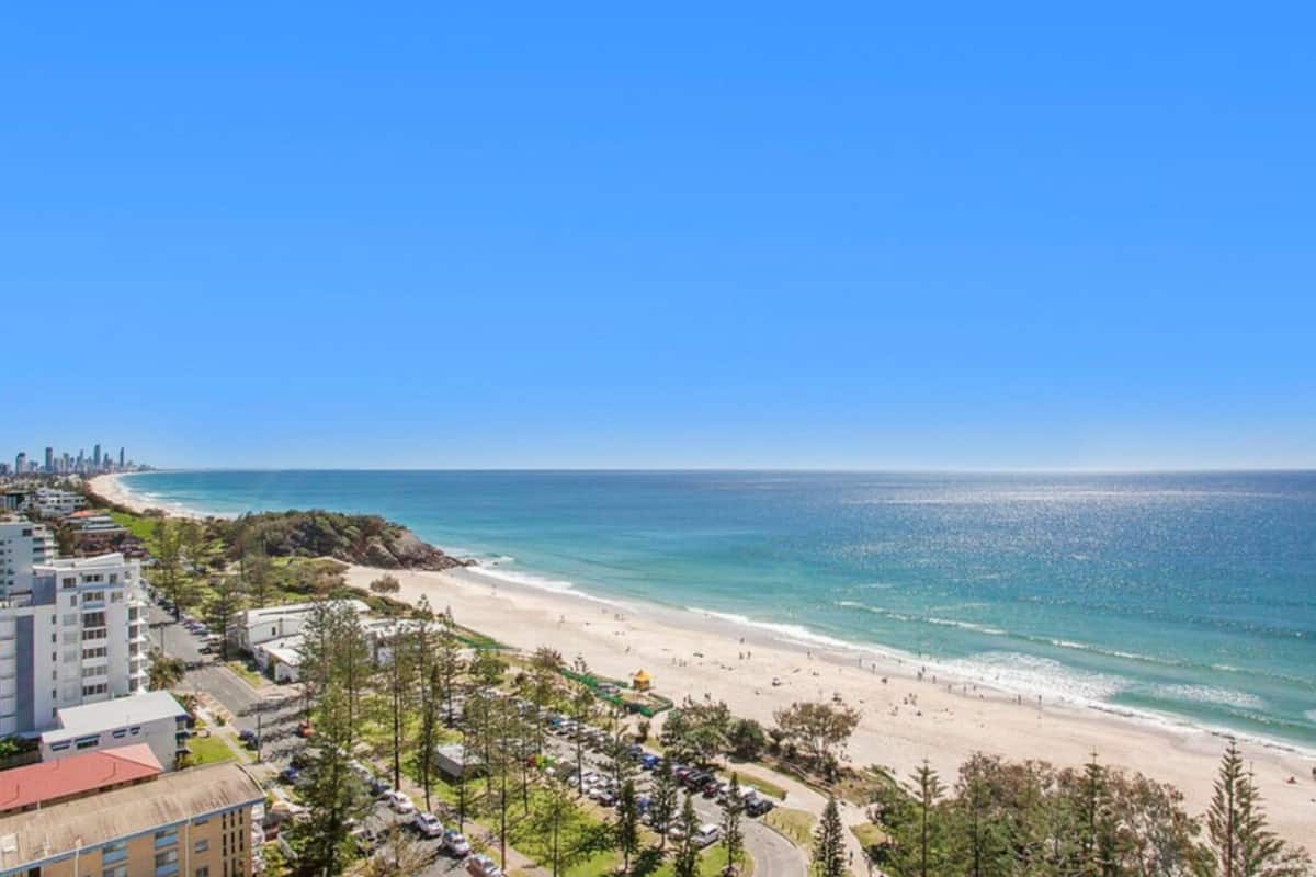A coastal hotel with a beach view, a parking lot, and a city skyline in the distance.