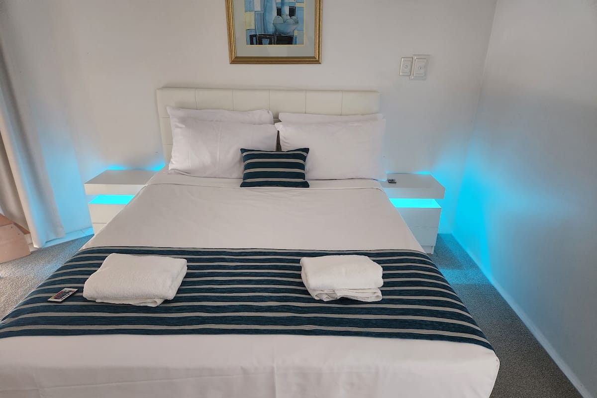 A neatly made bed with white and striped bedding, two white pillows, and two towels. There is a bedside table with a phone and a small lamp. The room has a simple, clean design with a framed picture on the wall.