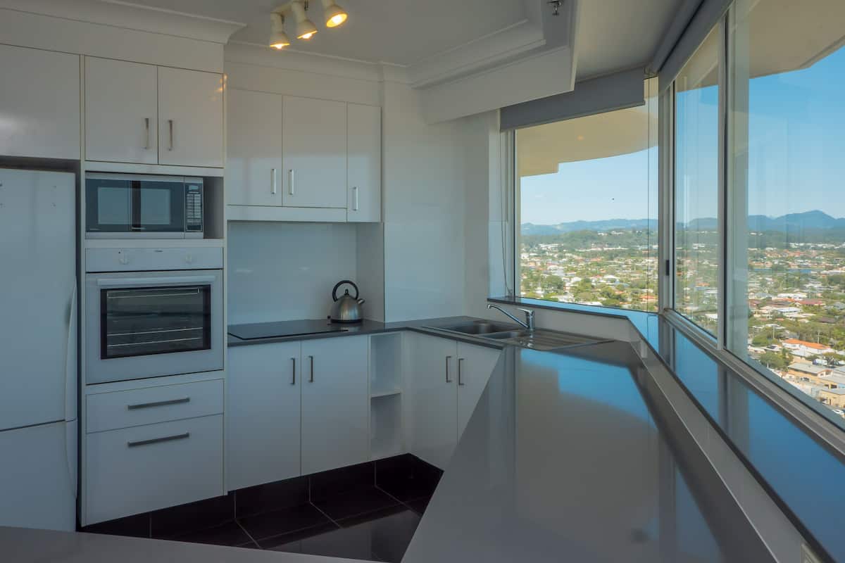 A modern kitchen with white cabinets, a microwave, an oven, and a kettle. It features a large window with a view of a cityscape.