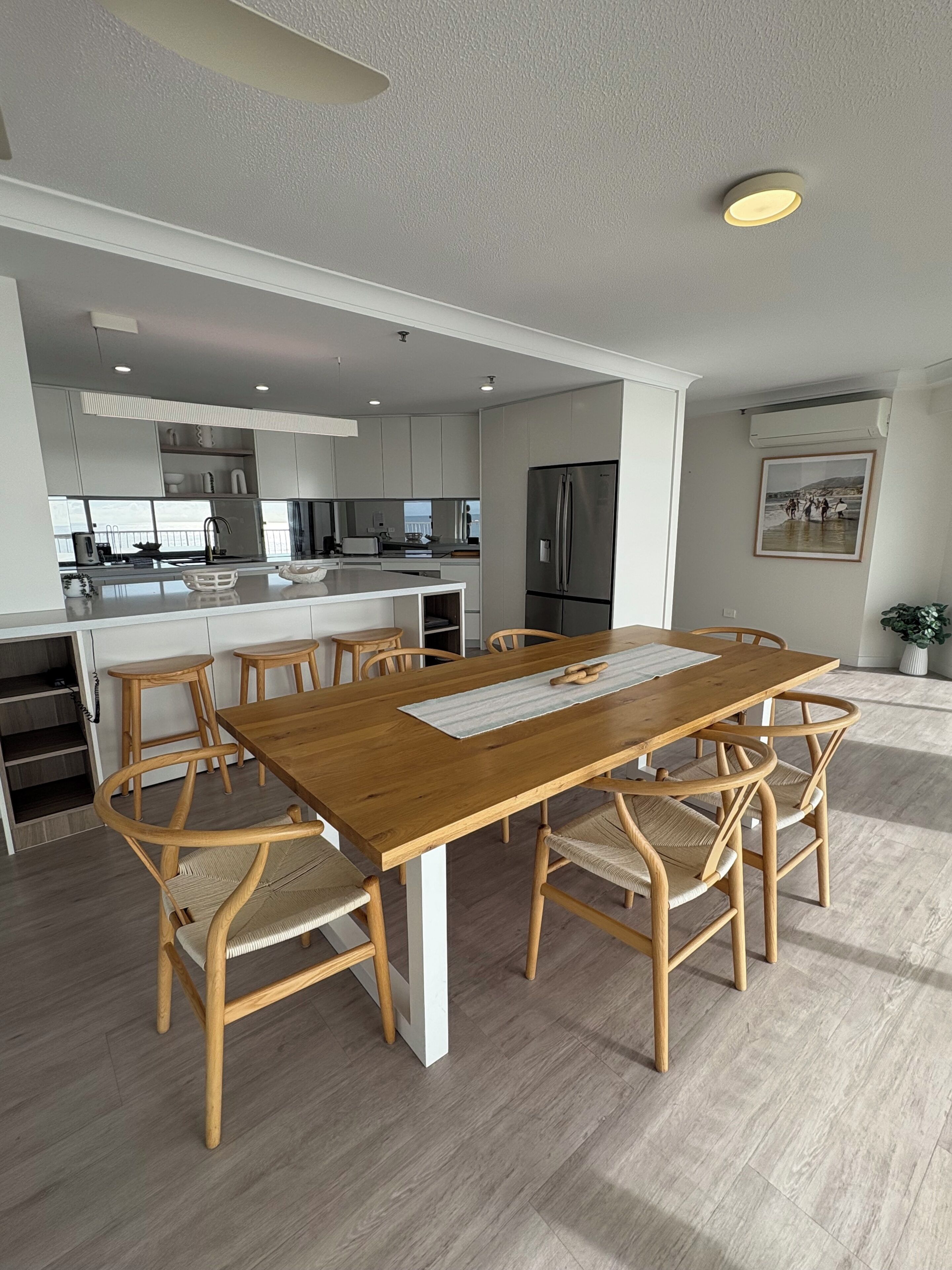 Una cocina moderna con armarios blancos, un refrigerador de acero inoxidable y un comedor con mesa y sillas de madera.