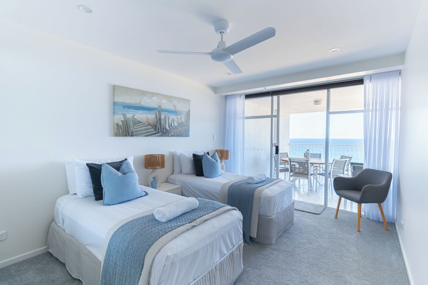 A hotel room with two beds, a ceiling fan, a painting, a balcony with a view of the sea, and a chair.